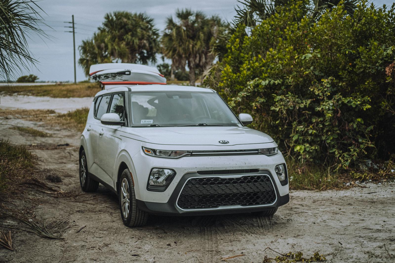 A white Kia Soul X-Line parked on a dirt road surrounded by lush greenery, featuring a roof cargo box.