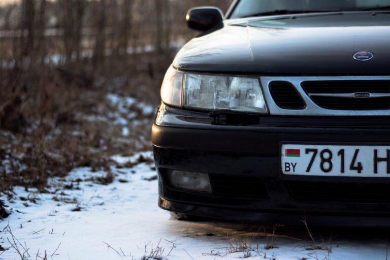 Close-up of a black car on a snowy terrain with focus on the headlight and license plate.