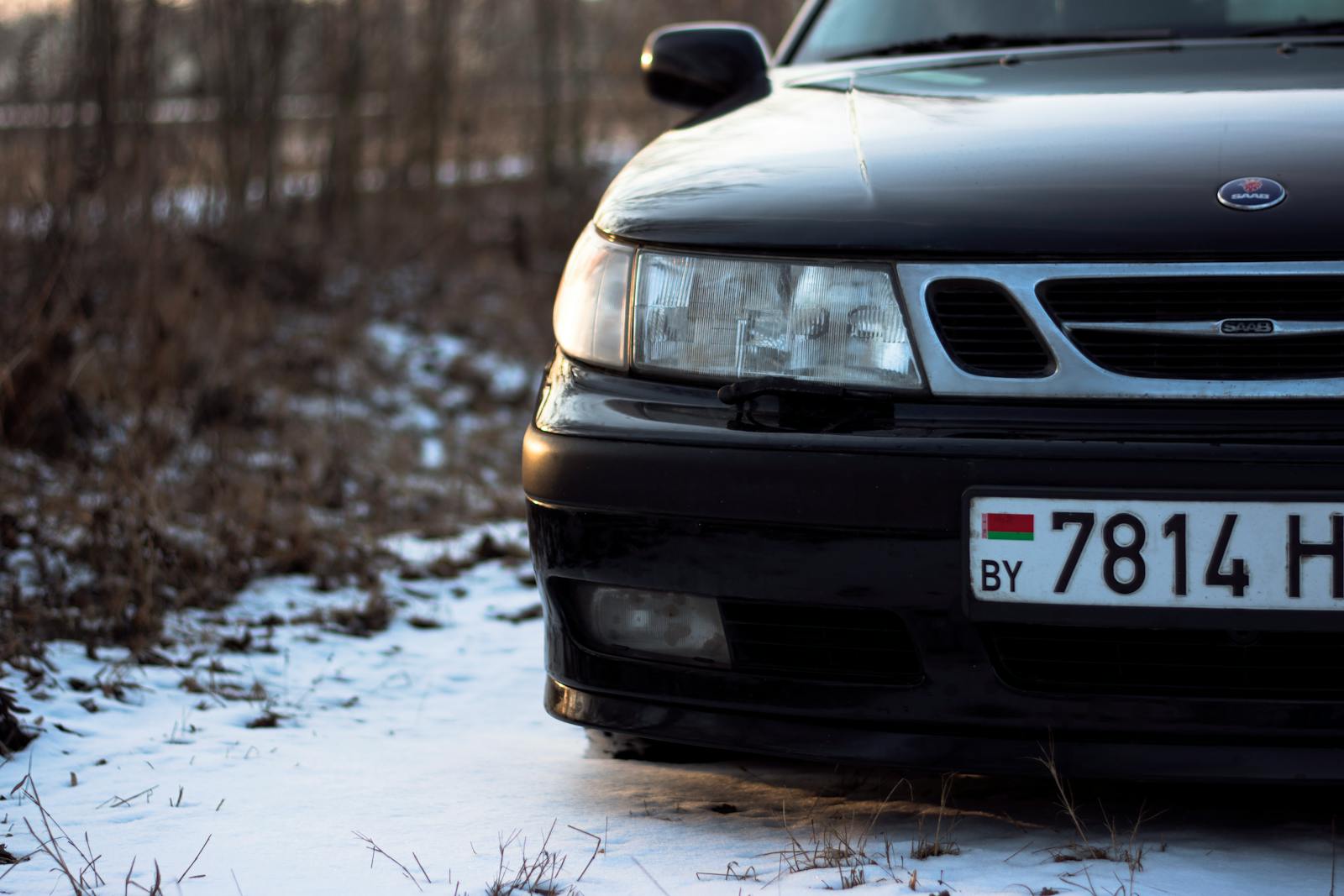 Close-up of a black car on a snowy terrain with focus on the headlight and license plate.