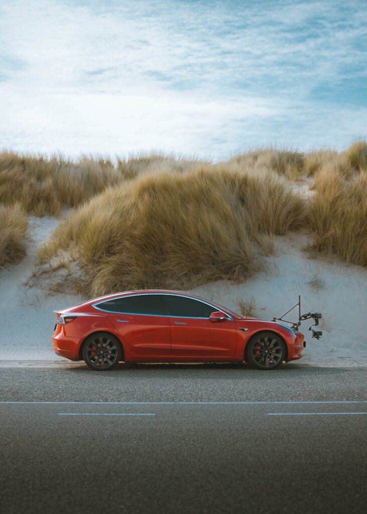 A sleek red electric car parked on a deserted road with grass dunes under a clear sky.