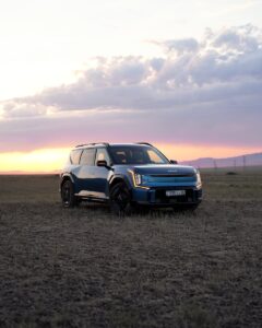 A blue electric SUV parked in a grassy field during sunset with dramatic sky and canyon backdrop.