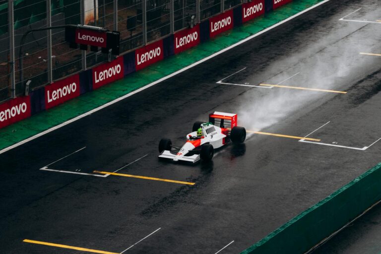 A classic Formula 1 McLaren car races on a wet track at Interlagos, Brazil, leaving a trail of spray behind.